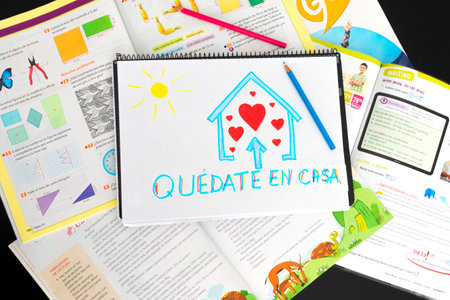 Benidorm, Spain - March 16, 2020 - Coronavirus pandemic self quarantine in Spain. Kid drawing picture with spanish words Quedate en casa - Stay at home - over school textbooks on tableのeditorial素材