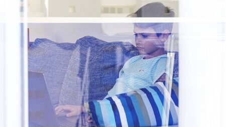 Child learning with laptop computer at home. View through the window. Technology, online learning,...
