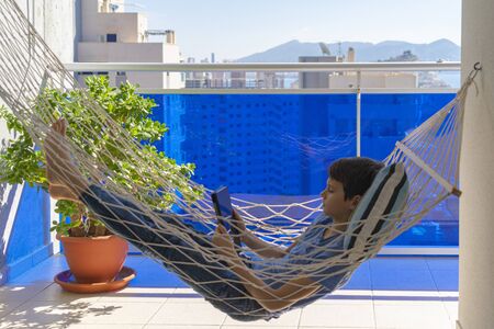 Boy with digital tablet computer lying in the hammock hanging on balcony. Education, online distance learning, homeschooling for kidsの写真素材