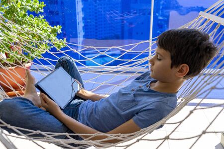 Boy with digital tablet computer lying in the hammock hanging on balcony. Education, online distance learning, homeschooling for kidsの写真素材