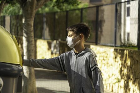 Child with protective mask and gloves throwing empty plastic bottle into recycling bin near...