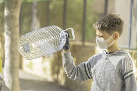 Child with protective mask and gloves throwing empty plastic bottle into recycling bin near residential apartment building outdoorsの写真素材