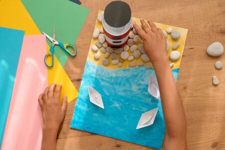 Kid hands make a lighthouse for school project at home. Top down view to wooden table with colored...