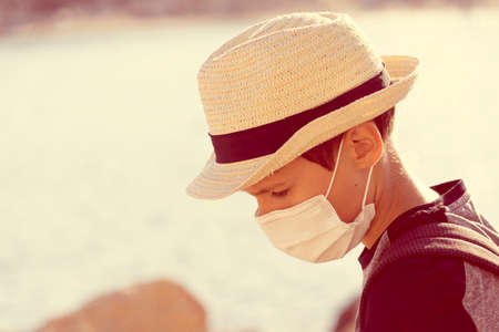 Boy with sun fedora hat and protective mask standing lowered head outdoors. Vacation, holiday, travel conceptの写真素材