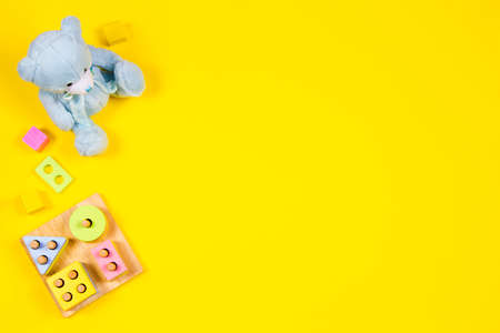 Baby kids toys background. Blue teddy bear, wooden educational stacking color recognition puzzle toy and colorful blocks on yellow background. Top view, flat layの写真素材