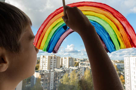 Boy painting colorful rainbow on window during Covid-19 quarantine at home. Stay at home due to danger of coronavirus infection. Symbol of hopeの写真素材
