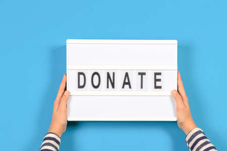 Hands holding light box with Donate word text over light blue background. Top view