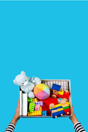 Donation concept. Kid hands with donate box with clothes, books and toys on light blue background....