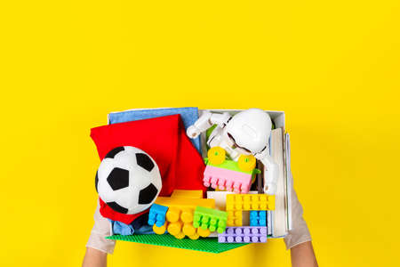 Donation concept. Woman hands with gloves holding donate box with clothes, books and toys on yellow...