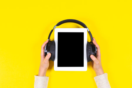 Online learning, remote education, e-learning concept. Kid hands holding digital tablet with wireless headphones over yellow background. Top viewの写真素材