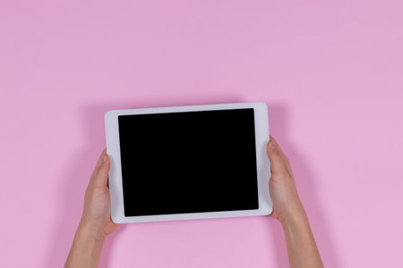 Kid hands holding digital tablet computer on pastel pink background. Top view