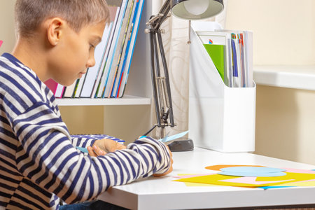 Education, learning, paper craft, entertainment. Boy cuts colored paper with scissors and making craft project at homeの写真素材