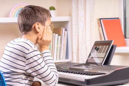 Teenage boy watching video lesson at tablet computer and learing playing digital piano at home. Online learning remote educationの写真素材