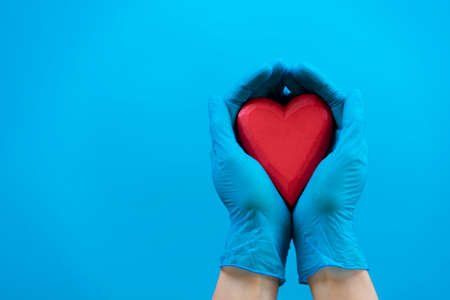 Hands wearing blue medical gloves holding red heart over light blue background. Health care, heart...