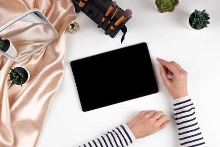 Female hands using digital tablet computer. Make up cosmetics, brushes on white desk. Top viewの写真素材