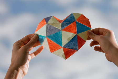 Female adult and child hands holding colorful paper heart over blue sky background. Autism spectrum...