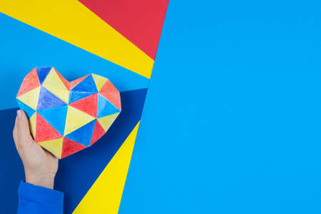 Child hand holding colorful paper heart over multicolored paper background. World autism awareness...
