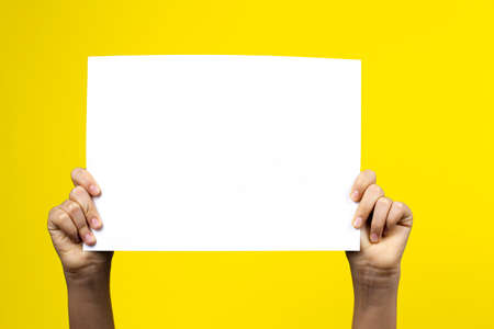Hands holding blank white paper board poster placard on yellow backgroundの写真素材