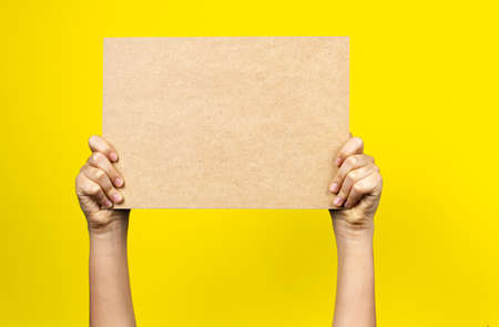 Hands holding blank brown paper cardboard board poster on yellow backgroundの写真素材