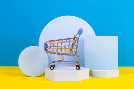 Mini shopping trolley cart with geometric podium platforms on yellow and light blue background. Online shopping, supermarket, store, sale conceptの写真素材
