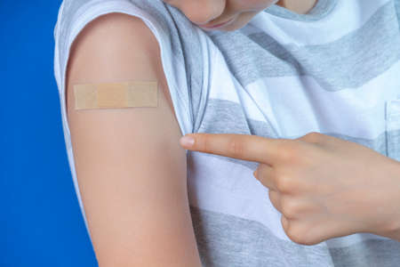 Teenage boy show to adhesive bandage plaster on his arm after vaccination on blue background. Injection covid vaccine, healthcare for children and teenagersの写真素材