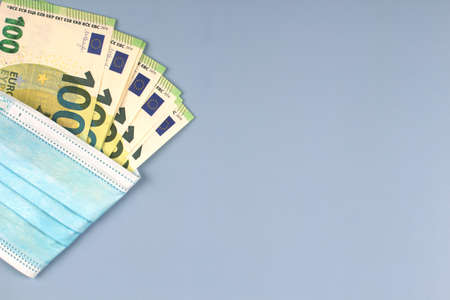 Some one hundred euro banknotes wrapped in disposable protective surgical face mask on light gray background. Top viewの写真素材