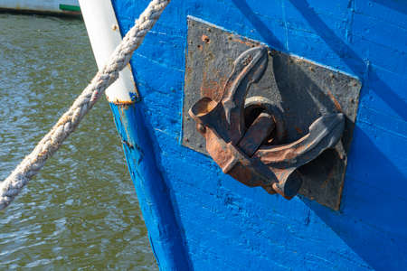 Old rusty ship anchor in up position on moored blue small boat. Close upの写真素材