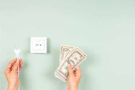 Woman hands holding electric power plug and US dollar banknotes near white electric socket on light green background. Electricity cost and expensive energy conceptの写真素材