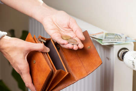 Womans hand takes euro money from wallet and places on heating radiator battery with temperature regulator. Expensive heating costs during cold season. Paying billsの写真素材