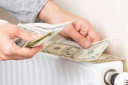 Womans hand counting US dollars banknotes and place on heating radiator battery with temperature...
