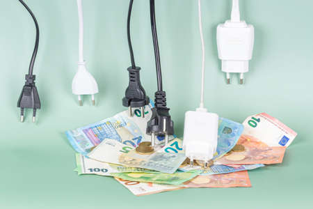 Many power cable cords with Eu plugs hanging over pile of Euro money coins and bills. Energy efficiency, power consumption, electricity cost, and expensive energy conceptの写真素材