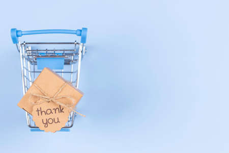 Top view to mini shopping cart on light blue background. Kraft paper box with tag inscription Thank you inside trolley. E-commerce, online shopping conceptの写真素材