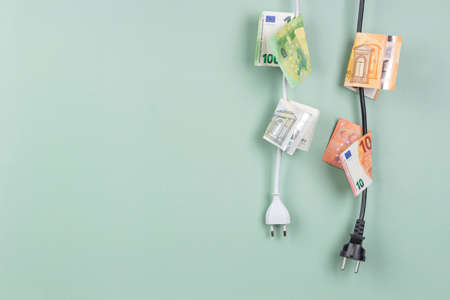 Electric power plugs with Euro banknotes on them hanging on light green background. Energy efficiency, power consumption, electricity cost, and expensive energy conceptの写真素材