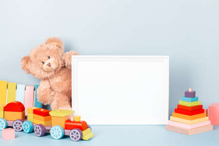 White wooden picture frame with blank mock up copy space standing next to teddy bear and colorful wooden toy train and stacking pyramid on light blue background. Front viewの写真素材
