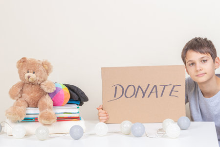 Charity, donation. Smiling kid holding brown piece of cardboard with inscription Donate. Volunteer collect kid clothes, books and toys for donation.の写真素材