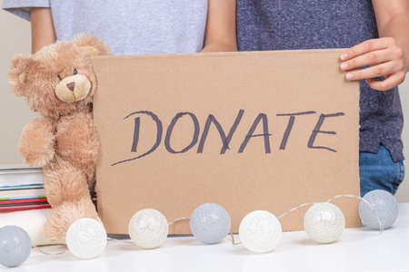Volunteer hands holding brown piece of cardboard with inscription Donate. Kids collect clothes,...