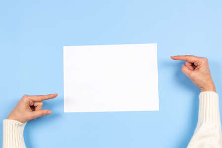 Female hands pointing to blank white paper sheet on light blue background. Top view. Mockup paper...