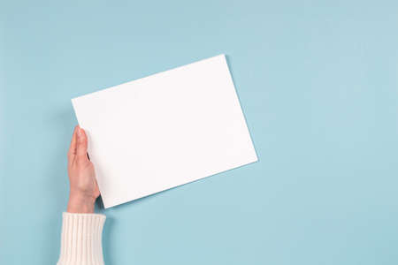 Female hands holding blank white paper sheet on light blue background. Top view. Mockup paper with...