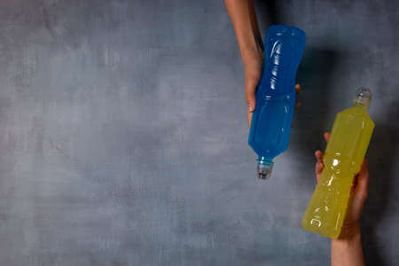 Hands holding plastic bottles with isotonic energy blue and yellow transparent sport drink on grungy gray background. Top view, copy space for textの写真素材