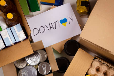 Donation for people from Ukraine. Food donations boxes with grocery products on white desks. Top...