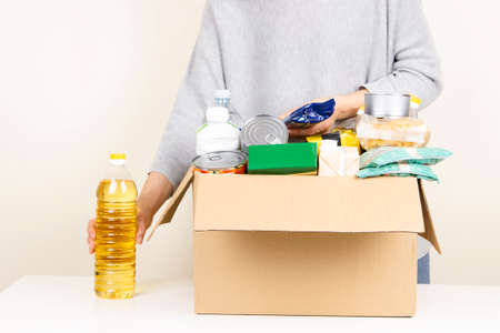 Volunteer hands collecting food into donation box full of grocery products. Donation, charity, food...