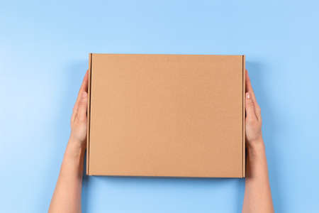 Top view to female hands holding brown cardboard box on light blue background. Mockup parcel box....