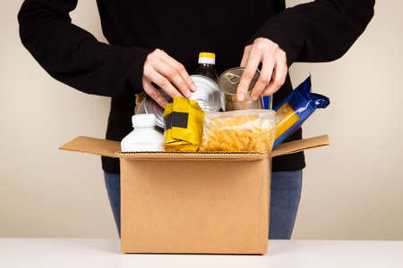 Volunteer hands collecting food into donation box. Cardbox full of grocery products. Donation, charity, food bank, help for poor families, migrants, refugeesの写真素材