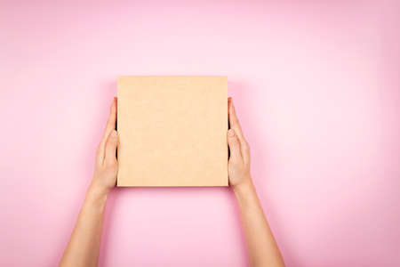 Top view to female hands holding brown cardboard box on pastel pink background. Mockup parcel box. Packaging, shopping, delivery conceptの写真素材