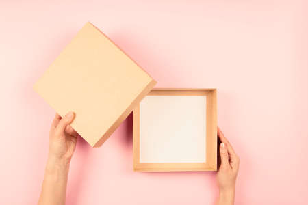 Female hands open empty brown cardboard box on light pink background. Mockup parcel box. Top view,...