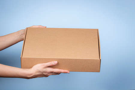 Woman hands holding brown ecological package box made of natural corrugated cardboard. Mockup mailing parcel box on blue background. Packaging, shopping, delivery conceptの写真素材