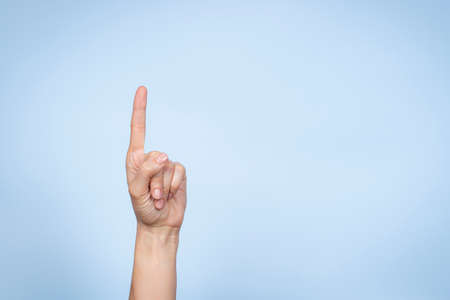 Hand gesture. Female hand shows number one. Woman pointing up with index finger on light blue backgroundの写真素材