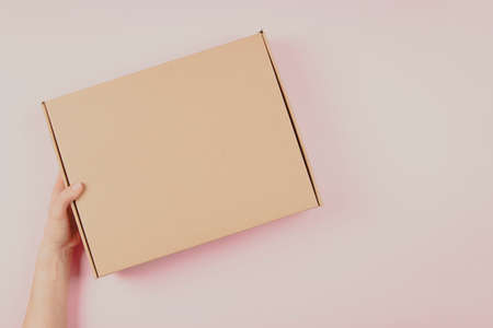 Female hand holding brown cardboard box on pastel pink background. Top view to mockup parcel box. Packaging, shopping, delivery conceptの写真素材