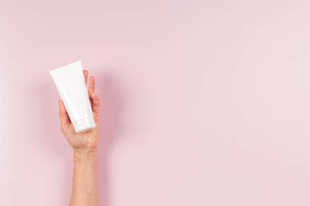 Woman hand holding white blank plastic cosmetics cream tube on pastel pink background. Skincare, moisturizer, face, body, hand cream, shampoo package container mock up with place for textの写真素材