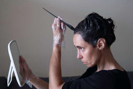 Middle age woman colouring dark hair with gray roots at home. Woman sitting on couch and dyeing hair using black brush and looking to the mirrowの写真素材